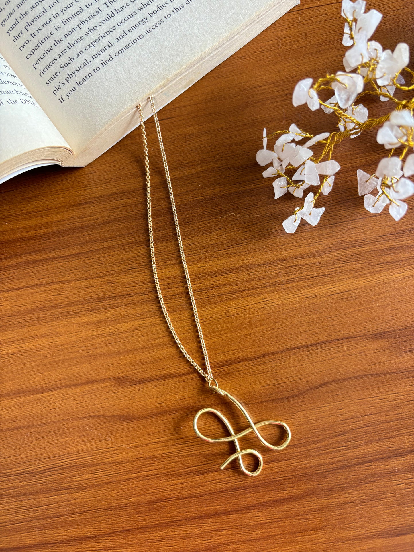 A gold-colored infinity pendant necklace displayed on a wooden surface next to a pair of white flower decorations and an open book.